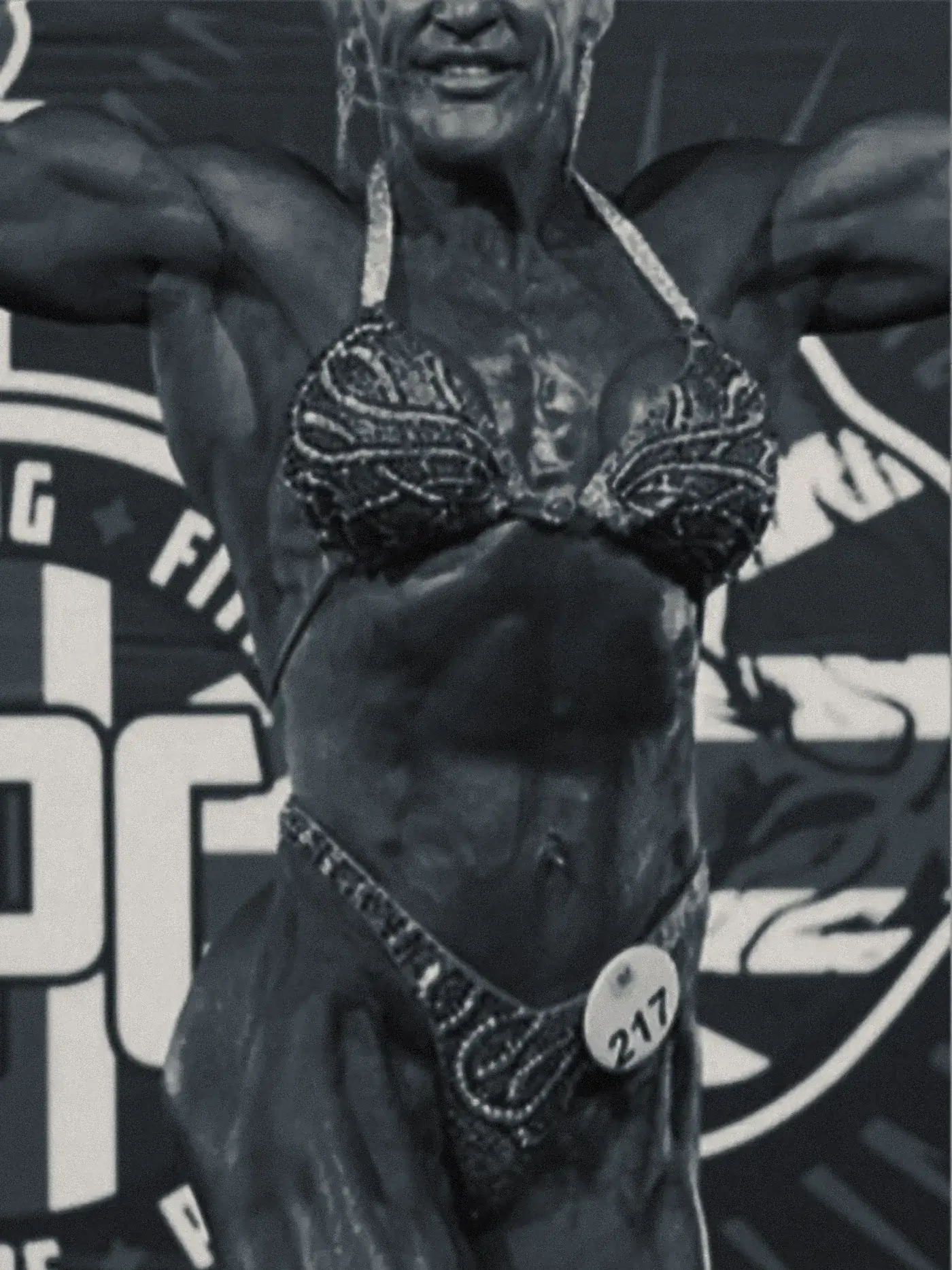 Julie on stage in full competition condition, holding a front pose under the lights